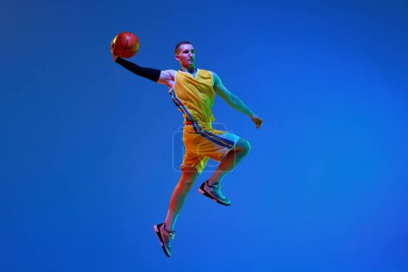 Téléchargez les photos : Jeune homme musclé en uniforme jaune, joueur de basket-ball pendant le match, sautant avec une balle contre fond bleu studio sous la lumière du néon. Concept de sport professionnel, passe-temps, mode de vie sain, action - en image libre de droit
