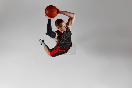 Photo for Dynamic image of concentrated young man, basketball player throwing all in a jump against grey studio background. Aerial view. Slam dunk. Concept of professional sport, competition, action and motion - Royalty Free Image