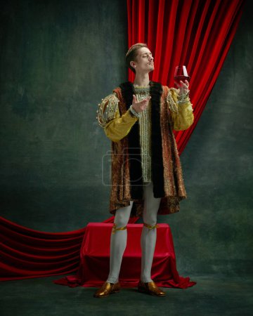 Téléchargez les photos : Portrait de jeune duc, prince, personne royale en costume vintage levant verre de vin rouge sur fond vert foncé, vintage. Concept de comparaison des époques, histoire, art de la renaissance, remake - en image libre de droit
