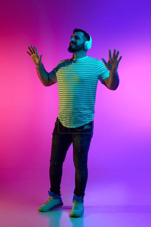 Photo for Full-length portrait of smiling bearded man in striped shirt listening to music in headphones against gradient pink blue studio background in neon light. Concept of human emotions, facial expression - Royalty Free Image