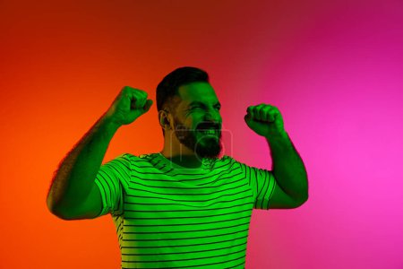 Photo for Portrait of happy bearded man posing with gesture of win and success against gradient studio background in neon light. Concept of human emotions, facial expression, lifestyle, business, promotion - Royalty Free Image