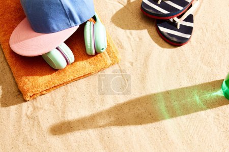 Photo for Shadow of beer bottle on warm sand with slippers, headphones, towel and cap. Summertime relaxation. Concept of alcohol drink, taste, summer vacation, holiday, brewery. Advertisement - Royalty Free Image