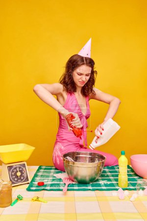 Foto de Chica joven concentrada e irritada en pastel de cocina vestido rosa para su cumpleaños contra fondo amarillo vivo. Concepto de fiesta, celebración, emociones, belleza femenina, juventud. Arte pop - Imagen libre de derechos