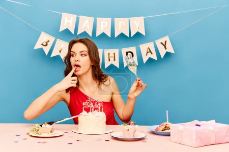 Photo for Beautiful young girl celebrating birthday, eating cake, drinking champagne over blue background. Pretty female model with bright makeup. Concept of party, celebration, emotions, female beauty, youth - Royalty Free Image