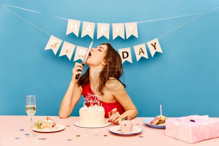 Photo for Beautiful, expressive young girl celebrating birthday, eating cake from knife against blue background. Concept of party, celebration, emotions, female beauty, youth. Pop art - Royalty Free Image