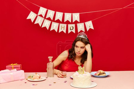 Téléchargez les photos : Jeune fille ennuyée et sans émotion assise à table, buvant, fêtant son anniversaire sur fond rouge. Vieillir. Concept de fête, célébration, émotions, beauté féminine, jeunesse. Pop art - en image libre de droit