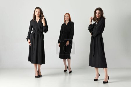 Foto de Retrato de tres estiletes, hermosas mujeres jóvenes en elegantes vestidos negros posando sobre fondo de estudio de luz. Concepto de moda elegante, belleza femenina, negocios, compras, ventas, anuncios - Imagen libre de derechos