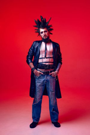 Photo for Portrait of young guy in image of punk with extraordinary hairstyle, makeup posing against red studio background in neon light. Concept of music, lifestyle, subculture, art, youth, human emotions - Royalty Free Image