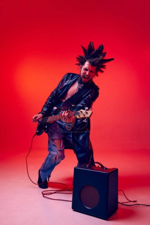 Téléchargez les photos : Jeune homme, punk en vêtements extraordinaires et coiffure jouant de la guitare électrique sur fond de studio rouge au néon. Concept de musique, style de vie, sous-culture, art, jeunesse, émotions humaines - en image libre de droit