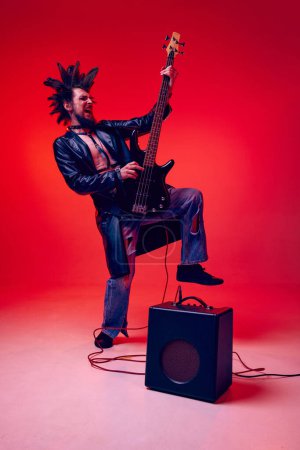 Photo for Rock musician. Young man, punk in extraordinary clothes and hairdo playing electric guitar against red studio background in neon light. Music, lifestyle, subculture, art, youth, human emotions concept - Royalty Free Image