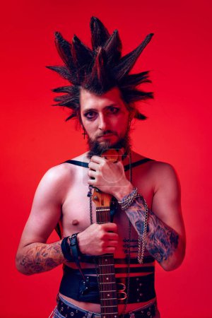 Photo for Deep look. Portrait of creative young man, punk, musician posing with electric guitar against red studio background in neon light. Concept of music, lifestyle, subculture, art, youth, human emotions - Royalty Free Image