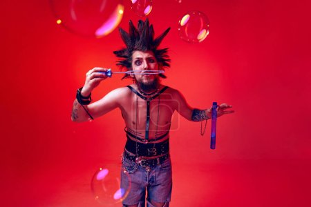 Photo for Crazy looking young man, punk, musician posing shirtless with soap bubbles against red studio background in neon light. Concept of music, lifestyle, subculture, art, youth, human emotions - Royalty Free Image