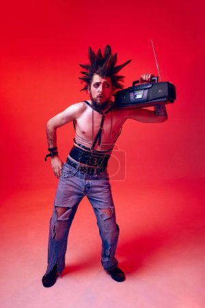 Photo for Young man, punk in extraordinary clothes and hair listening to music with retro player against red studio background in neon light. Concept of music, lifestyle, subculture, art, youth, human emotions - Royalty Free Image