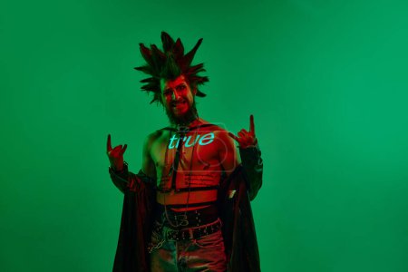 Téléchargez les photos : Jeune homme expressif, musicien rock, punk avec filtre au néon sur le corps posant sur fond de studio vert au néon. Concept de musique, style de vie, sous-culture, art, jeunesse, émotions humaines - en image libre de droit