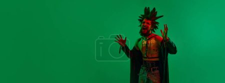 Photo for Emotional young man, punk with extraordinary hairstyle and makeup posing over green background in neon light. Concept of music, lifestyle, subculture, art, youth, emotions. Banner. Copy space for ad - Royalty Free Image