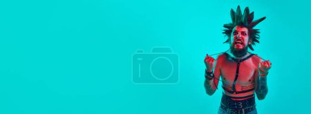 Photo for Shirtless young man, punk with crazy hairdo and makeup posing over blue studio background in neon light. Concept of music, lifestyle, subculture, art, youth, human emotions. Banner. Copy space for ad - Royalty Free Image