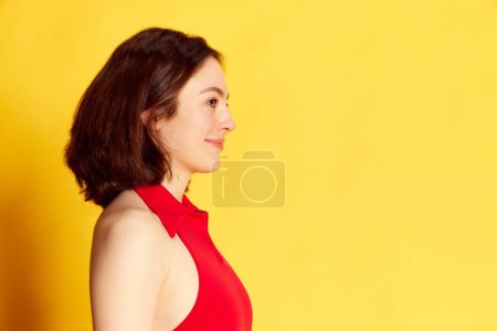 Téléchargez les photos : Vue du profil de la jeune femme brune en vêtements décontractés debout, posant, regardant droit sur fond de studio jaune. Concept de jeunesse, émotions humaines, style de vie, beauté, mode, publicité - en image libre de droit