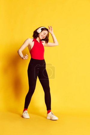 Retrato de larga duración de una joven hermosa vestida con ropa casual escuchando música en auriculares y bailando sobre fondo amarillo del estudio. Concepto de juventud, emociones humanas, estilo de vida, anuncio