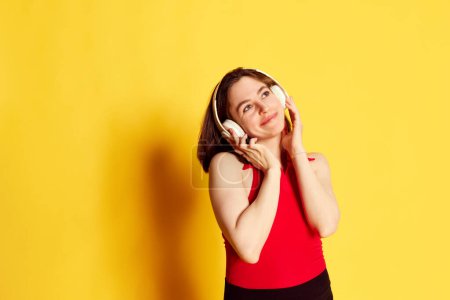 Foto de Retrato de una hermosa y atractiva joven morena escuchando música en auriculares sobre fondo de estudio amarillo. Concepto de juventud, emociones humanas, estilo de vida, belleza, anuncio - Imagen libre de derechos