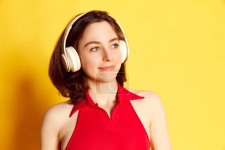 Téléchargez les photos : Portrait d'une belle jeune fille brune attrayante écoutant de la musique dans un casque sur fond de studio jaune. Concept de jeunesse, émotions humaines, mode de vie, beauté, publicité - en image libre de droit