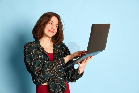 Photo for Portrait of young girl in checkered shirt working on laptop against blue studio background. Communication, business, education. Concept of youth, human emotions, lifestyle, ad - Royalty Free Image