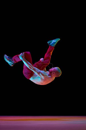 Téléchargez les photos : La pose du saut périlleux. Jeune homme artistique avec un fort corps musclé dansant breakdance sur fond de studio noir dans la lumière au néon. Concept d'art, danse de rue, mode, jeunesse, passe-temps, dynamique, publicité - en image libre de droit
