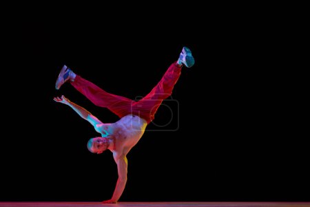 Téléchargez les photos : Tournage à la main. Jeune homme musclé, talentueux et actif dansant le breakdance sur fond de studio noir au néon. Concept d'art, danse de rue, mode, jeunesse, passe-temps, dynamique, publicité - en image libre de droit