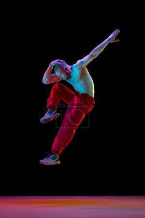 Téléchargez les photos : Jeune homme musclé avec relief corps torse nu dansant breakdance sur fond studio noir dans la lumière au néon. Concept d'art, danse de rue, mode, jeunesse, passe-temps, dynamique, publicité - en image libre de droit