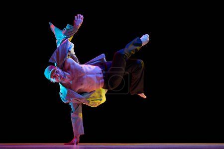 Téléchargez les photos : Jeune homme élégant en tenue décontractée dansant breakdance sur fond de studio noir au néon. Corps musculaire. Concept d'art, danse de rue, mode, jeunesse, passe-temps, dynamique, publicité - en image libre de droit