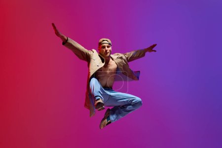 Téléchargez les photos : Jeune homme actif et artistique en tenue décontractée dansant breakdance, mépris contre fond studio violet rose. Concept d'art, danse de rue, mode, jeunesse, passe-temps, dynamique, publicité - en image libre de droit