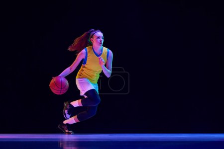 Photo for Young concentrated girl, basketball player in motion, running with ball against black studio background in neon light. Concept of professional sport, action and motion, game, competition, hobby, ad - Royalty Free Image