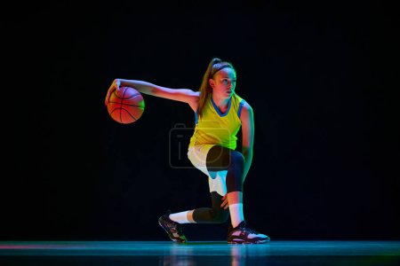 Photo for Young girl, professional basketball player in motion, training, playing against black studio background in neon light. Concept of professional sport, action and motion, game, competition, hobby, ad - Royalty Free Image