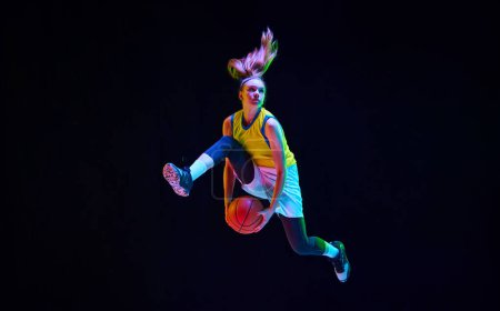 Photo for Dynamic image of motivated sportsman, young girl playing basketball, jumping on black studio background in neon light. Concept of professional sport, action and motion, game, competition, hobby, ad - Royalty Free Image