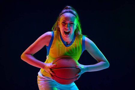 Photo for Concentrated and motivated young girl, basketball player in uniform posing with ball against black studio background in neon light. Concept of professional sport, action and motion, game, hobby, ad - Royalty Free Image