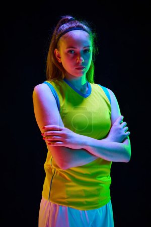 Photo for Portrait of young girl, basketball athlete posing in uniform with serious face against black background in neon light. Concept of professional sport, action and motion, game, competition, hobby, ad - Royalty Free Image