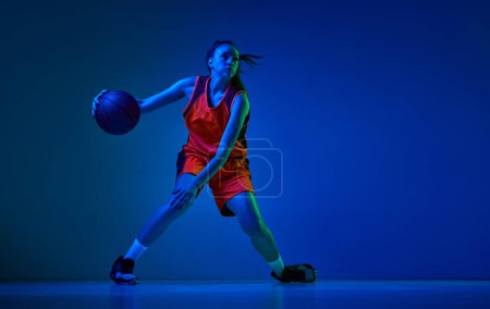 Photo for Dynamic image of young girl, basketball player in uniform in motion, playing over blue studio background in neon light. Concept of professional sport, action and motion, game, competition, hobby, ad - Royalty Free Image