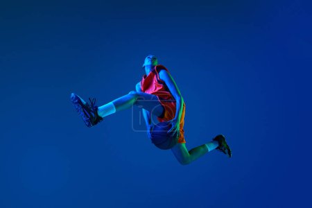 Foto de Vista inferior de la joven, jugador de baloncesto saltando con la pelota contra fondo estudio azul en luz de neón. Concepto de deporte profesional, acción y movimiento, juego, competición, hobby, anuncio - Imagen libre de derechos
