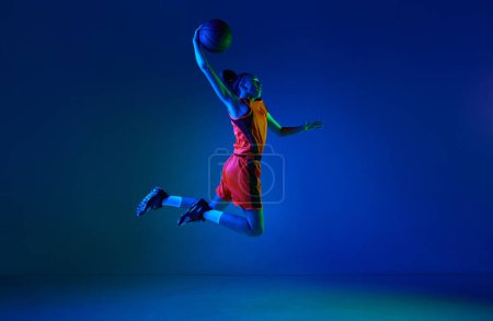 Photo for Slam dunk. Motivated young girl, basketball player throwing ball in jump against blue studio background in neon light. Concept of professional sport, action and motion, game, competition, hobby, ad - Royalty Free Image