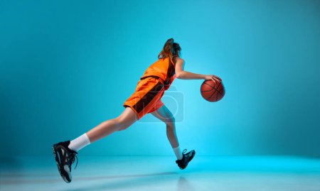 Foto de Vista posterior imagen dinámica de niña, jugador de baloncesto dribleando pelota contra fondo estudio azul en luz de neón. Concepto de deporte profesional, acción y movimiento, juego, competición, hobby, anuncio - Imagen libre de derechos