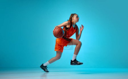 Photo for Young athletic girl, basketball player in motion, running with ball against blue studio background in neon light. Concept of professional sport, action and motion, game, competition, hobby, ad - Royalty Free Image