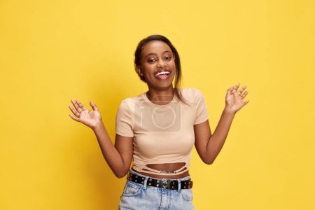 Téléchargez les photos : Portrait de belle, heureuse, jeune fille africaine en vêtements décontractés posant avec sourire et rire sur fond de studio jaune. Concept de style de vie, émotions humaines, mode, beauté, publicité - en image libre de droit