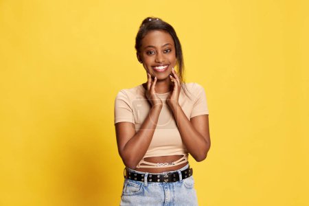 Téléchargez les photos : Portrait de belle, charmante, jeune fille africaine en vêtements décontractés posant avec un sourire tendre sur fond de studio jaune. Concept de style de vie, émotions humaines, mode, beauté, publicité - en image libre de droit