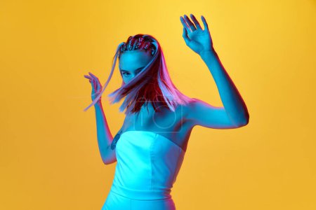 Photo for Portrait of young girl with pretty hairstyle dancing in white elegant dress against yellow studio background in neon light. Joy and fun. Concept of youth, emotions, beauty, lifestyle, ad - Royalty Free Image