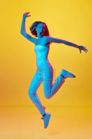Photo for Full-length portrait of young emotional girl in white clothes jumping with shocked face against yellow studio background in neon light. Concept of youth, emotions, beauty, lifestyle, ad - Royalty Free Image