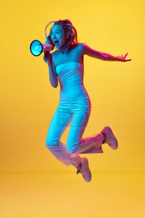 Photo for Full-length portrait of young emotional girl jumping and emotionally shouting megaphone against yellow studio background in neon light. Concept of youth, emotions, beauty, lifestyle, ad - Royalty Free Image