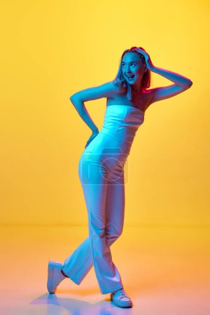 Photo for Full-length portrait of beautiful young girl in white clothes posing against yellow studio background in neon light. Concept of youth, emotions, beauty, fashion, lifestyle, ad - Royalty Free Image