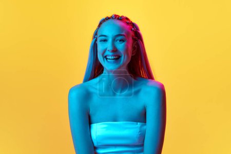 Photo for Portrait of beautiful young girl with pretty hairstyle looking at camera with smile and laughing against yellow studio background in neon light. Concept of youth, emotions, beauty, lifestyle, ad - Royalty Free Image