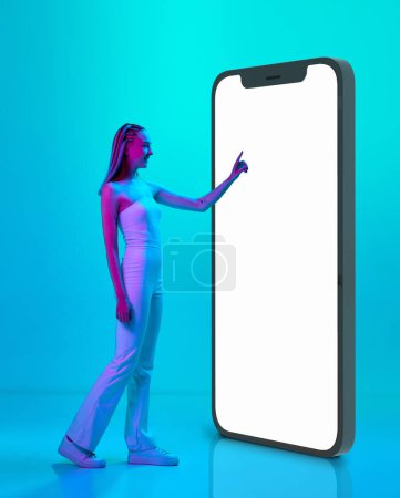 Photo for Online shopping. Young girl standing near giant 3D model of mobile phone screen against blue studio background in neon light. Concept of youth, emotions, beauty, lifestyle, ad - Royalty Free Image