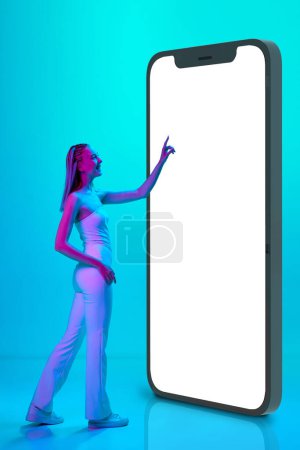 Foto de Compras en línea. Chica joven de pie cerca de gigante modelo 3D de la pantalla del teléfono móvil contra fondo de estudio azul en luz de neón. Concepto de juventud, emociones, belleza, estilo de vida, anuncio - Imagen libre de derechos
