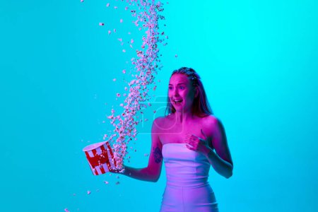 Photo for Horror movie. Young meotive girl with popcorn standing with expression of fear against blue studio background in neon light. Concept of youth, emotions, beauty, lifestyle, ad - Royalty Free Image